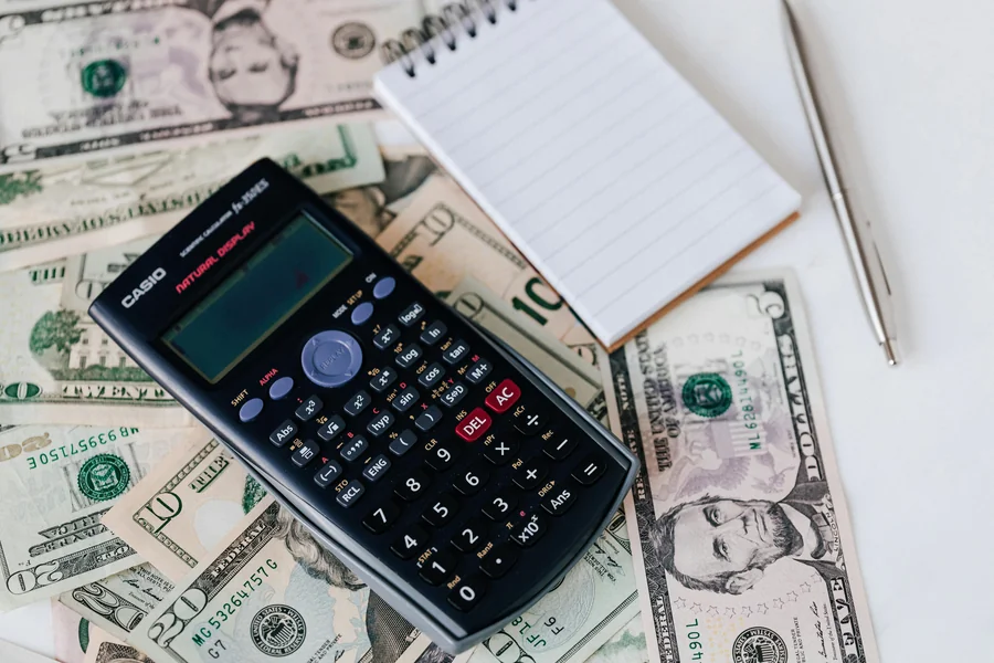 A calculator, notepad, and pen placed on top of US dollar bills representing financial planning and the real cost of starting over abroad